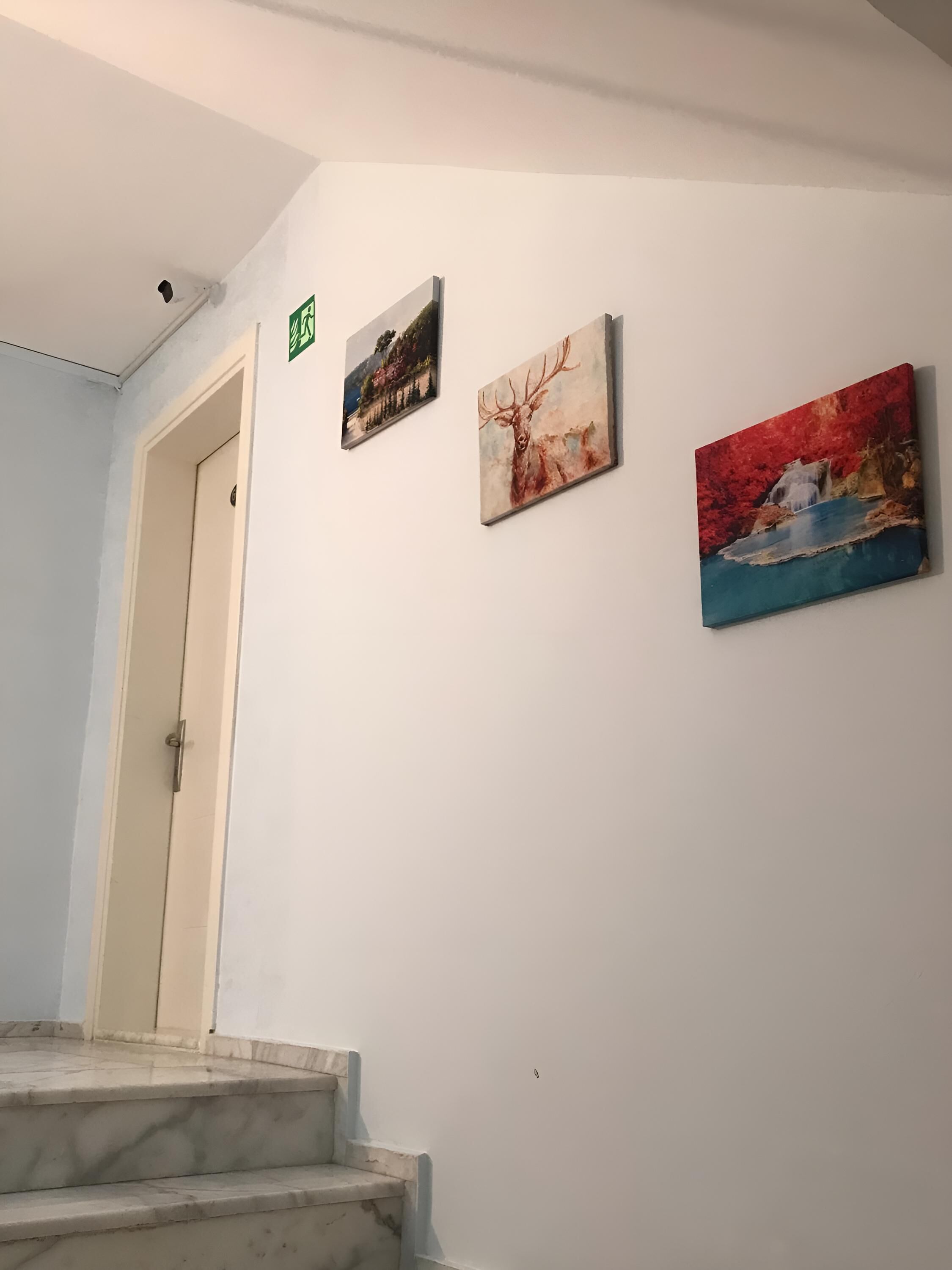 A hallway with a staircase leading to a door marked with an exit sign. Three paintings are displayed on the wall.