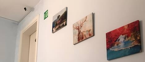 A hallway with a staircase leading to a door marked with an exit sign. Three paintings are displayed on the wall.