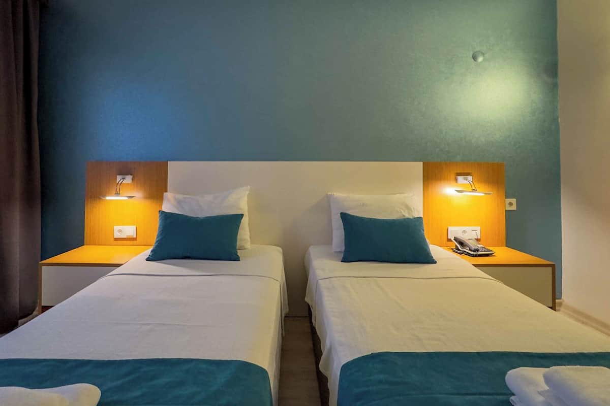 A hotel room with two single beds, each with a blue pillow and white linens. There are bedside lamps and a telephone on the nightstands.