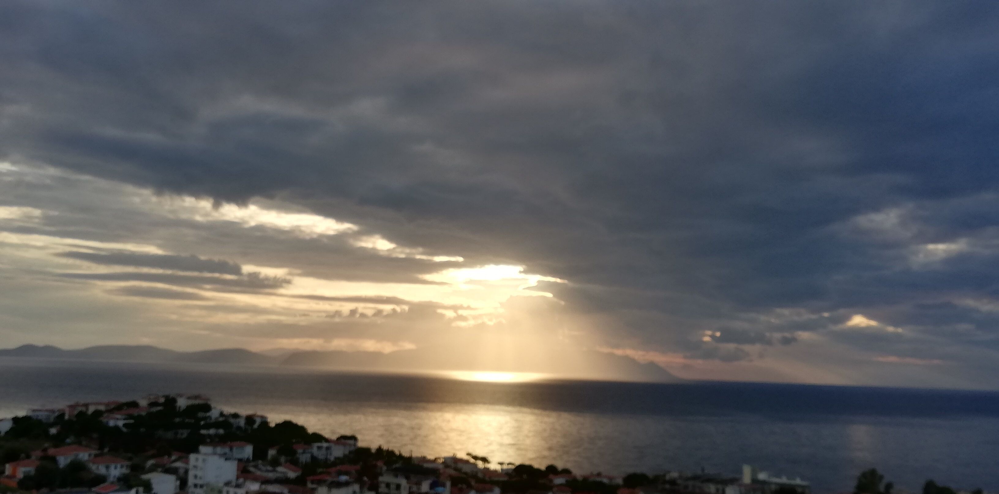 Un paisaje costero con un atardecer sobre el mar, un pueblo en primer plano y montañas a lo lejos.