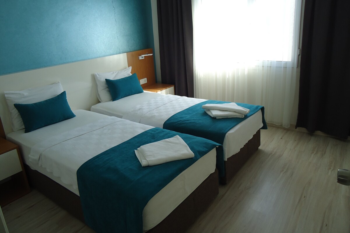 A hotel room with two beds, each with a blue bedspread and white pillows. There is a nightstand with a lamp between the beds, and a window with curtains on the right side.