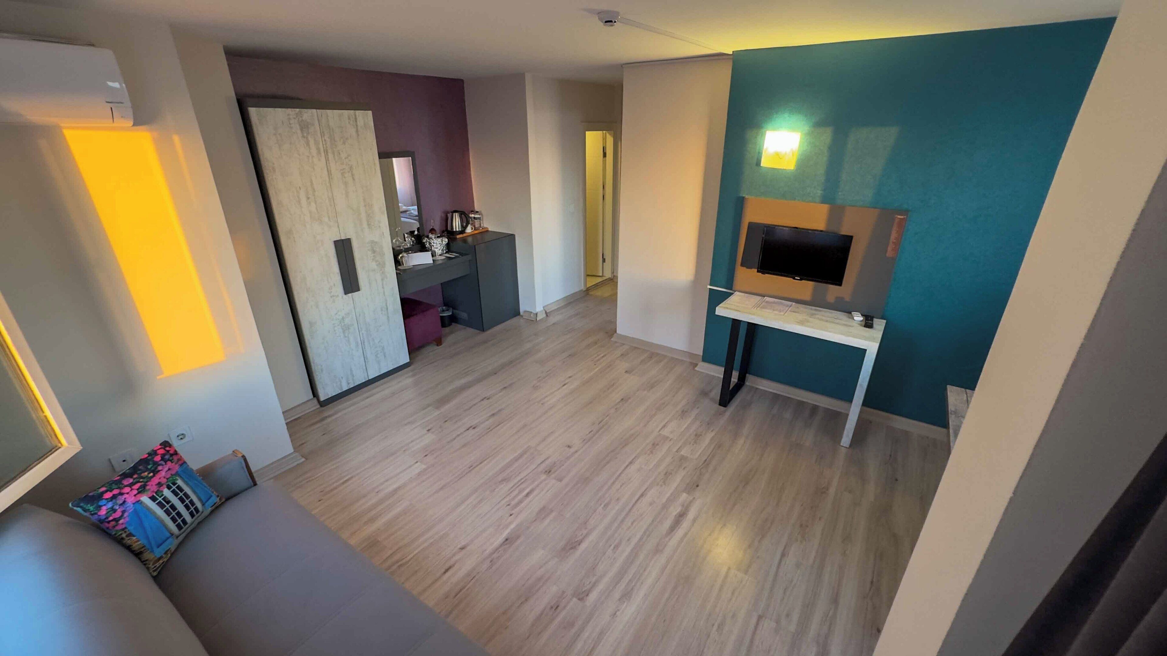Een moderne woonkamer met een houten vloer, een bank met een bloemenkussen, een tv op een dressoir en een deur die naar een andere kamer leidt.