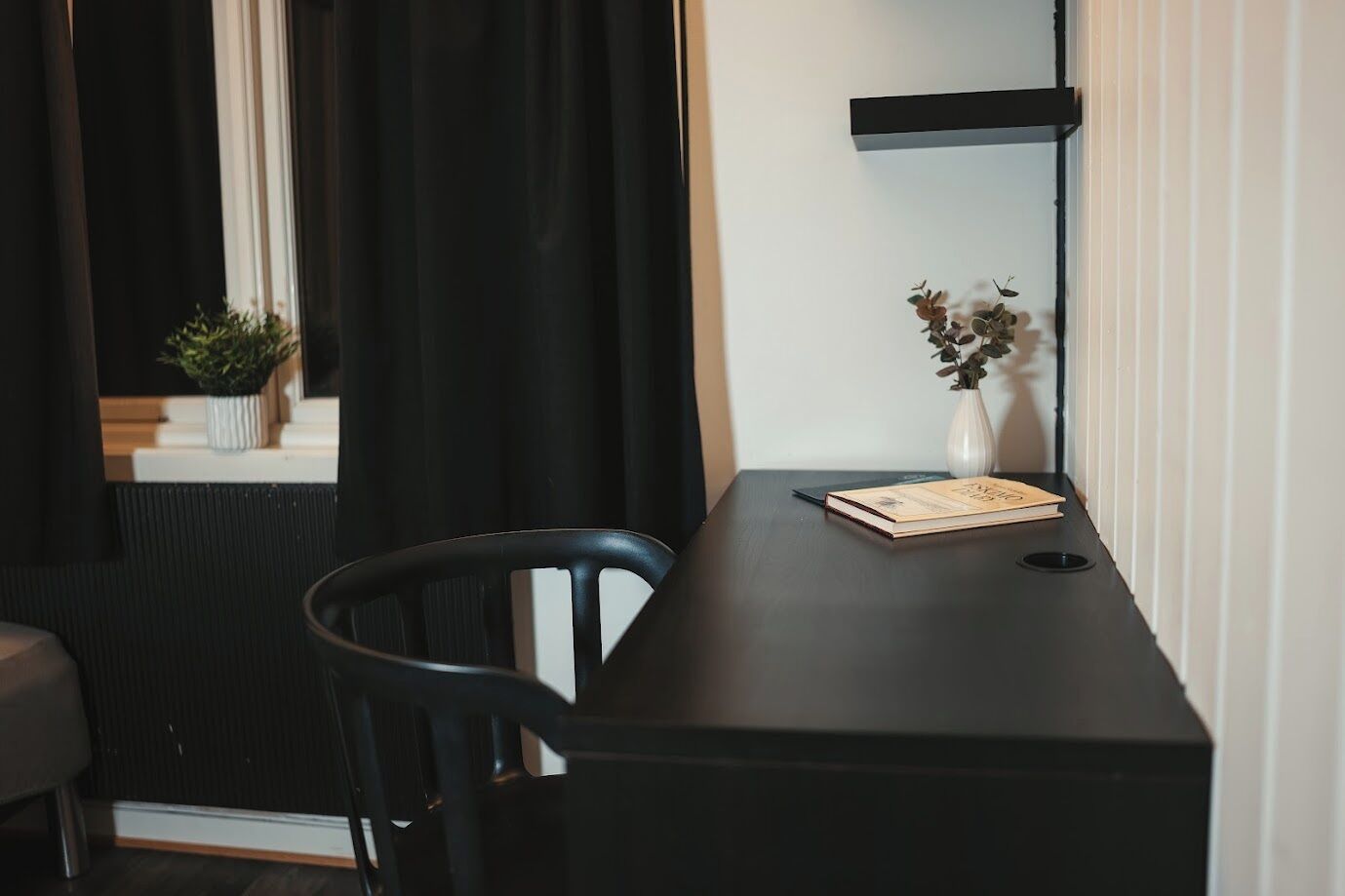A dark wooden desk with a book and a vase with dried flowers. A black chair is positioned in front of the desk.