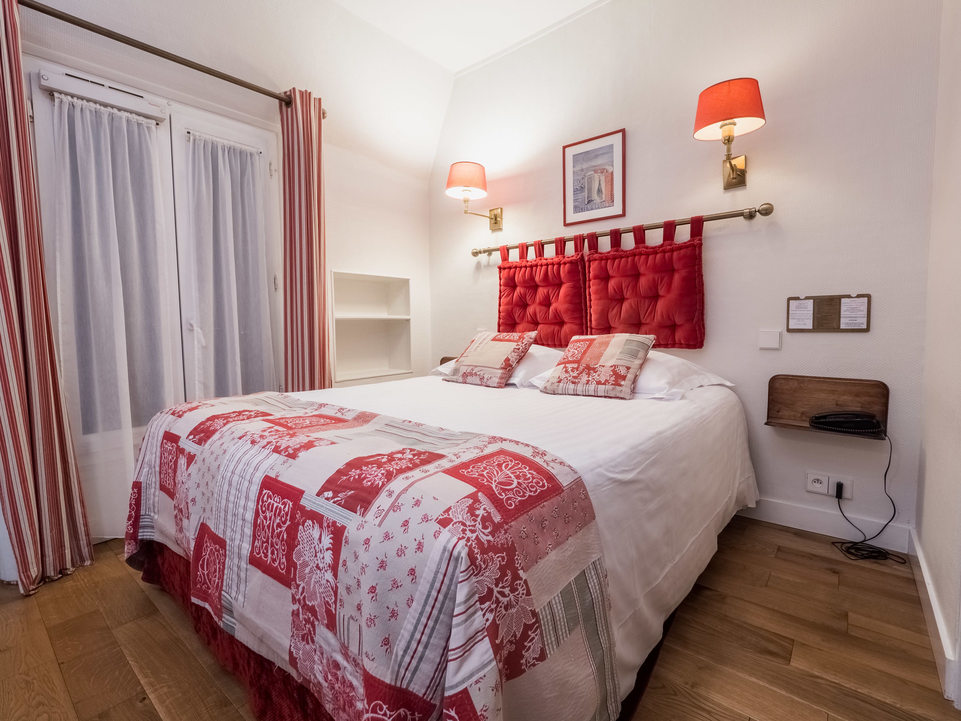 Una cama bien hecha con una colcha roja y blanca estampada, dos lámparas de pared, un cuadro enmarcado y un estante de madera con un teléfono.
