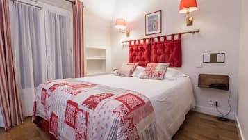 Una cama bien hecha con una colcha roja y blanca estampada, dos lámparas de pared, un cuadro enmarcado y un estante de madera con un teléfono.