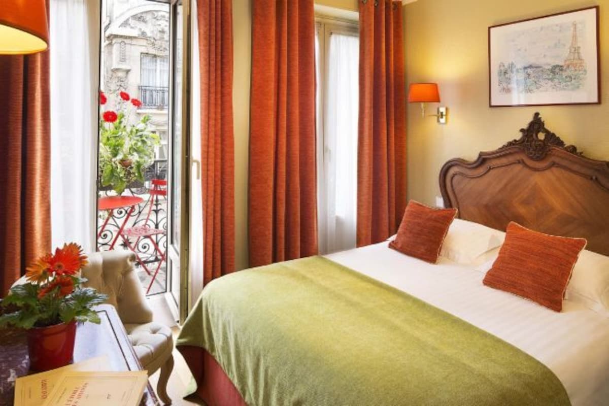 A hotel room with a large bed, a view of the street, and a balcony with flowers.