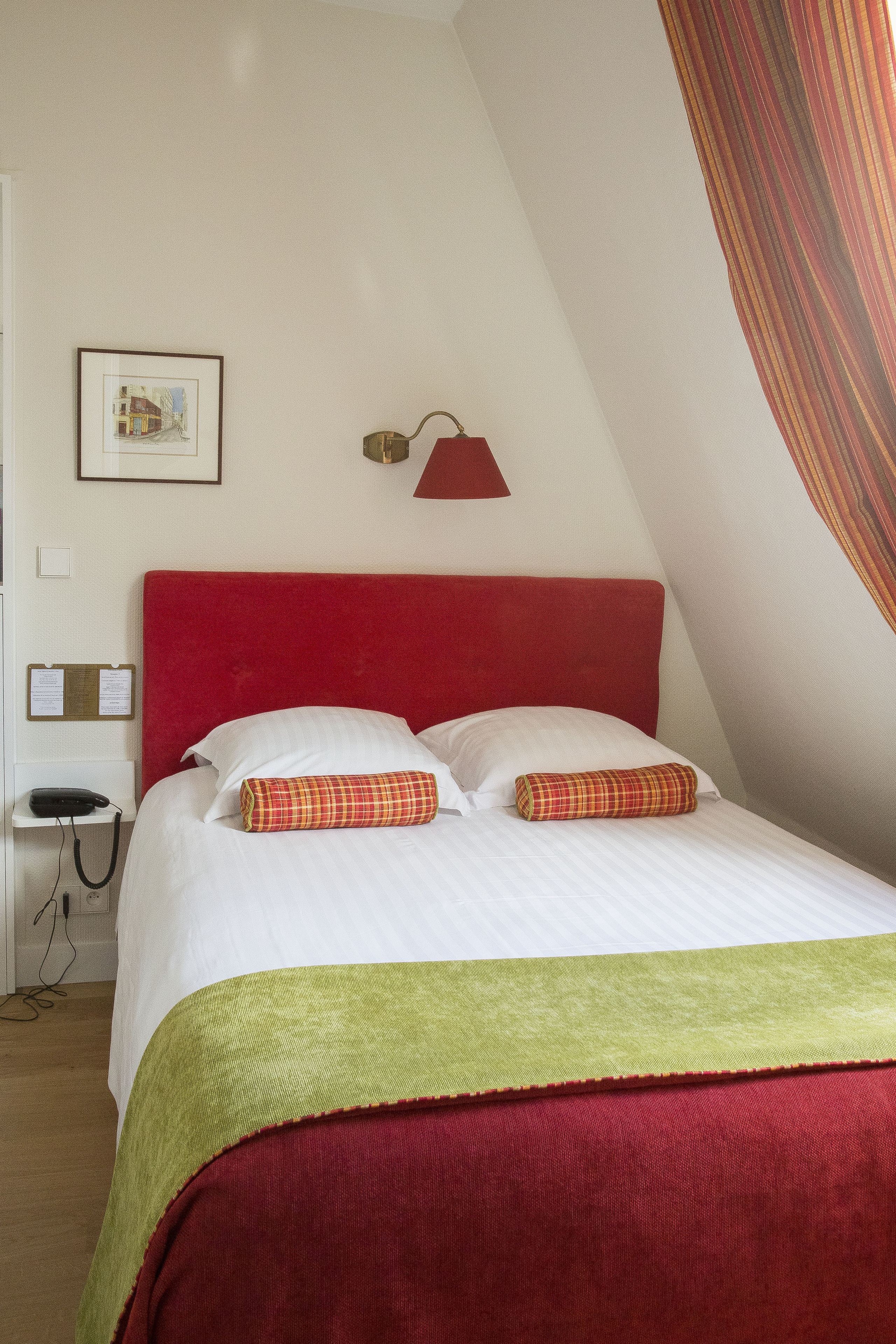 A neatly made bed with a red headboard, white bedding, and a green and red blanket. There is a framed picture on the wall and a red lamp above the bed.