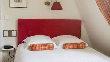 Un lit bien fait, avec une tête de lit rouge, du linge de lit blanc et une couverture verte et rouge. Un tableau encadré est accroché au mur et une lampe rouge est placée au-dessus du lit.