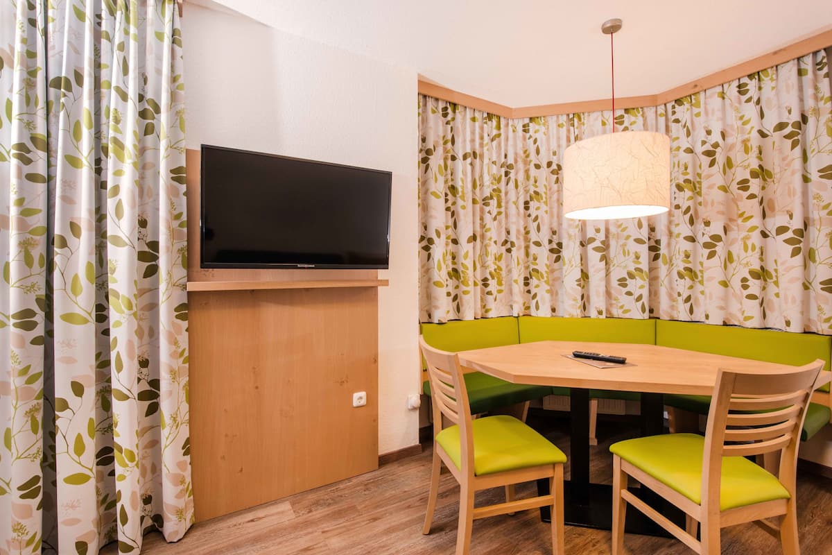 Ein Eckzimmer mit an einer Holdekoration befestigtem Fernseher, einem Tisch mit zwei Stühlen und einer grün gepolsterten Bank.