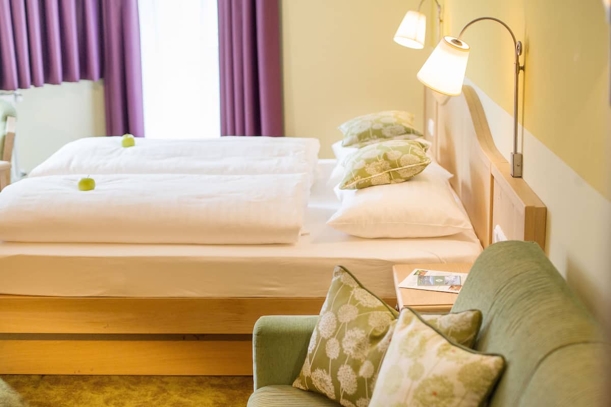 Ein Hotelzimmer mit einem Bett, zwei Kissen, einem Nachttisch mit Lampe und einem grünen Sofa mit floralen Kissen.