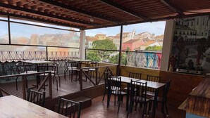 An outdoor dining area with wooden tables and chairs, a tiled floor, and a view of buildings in the background.