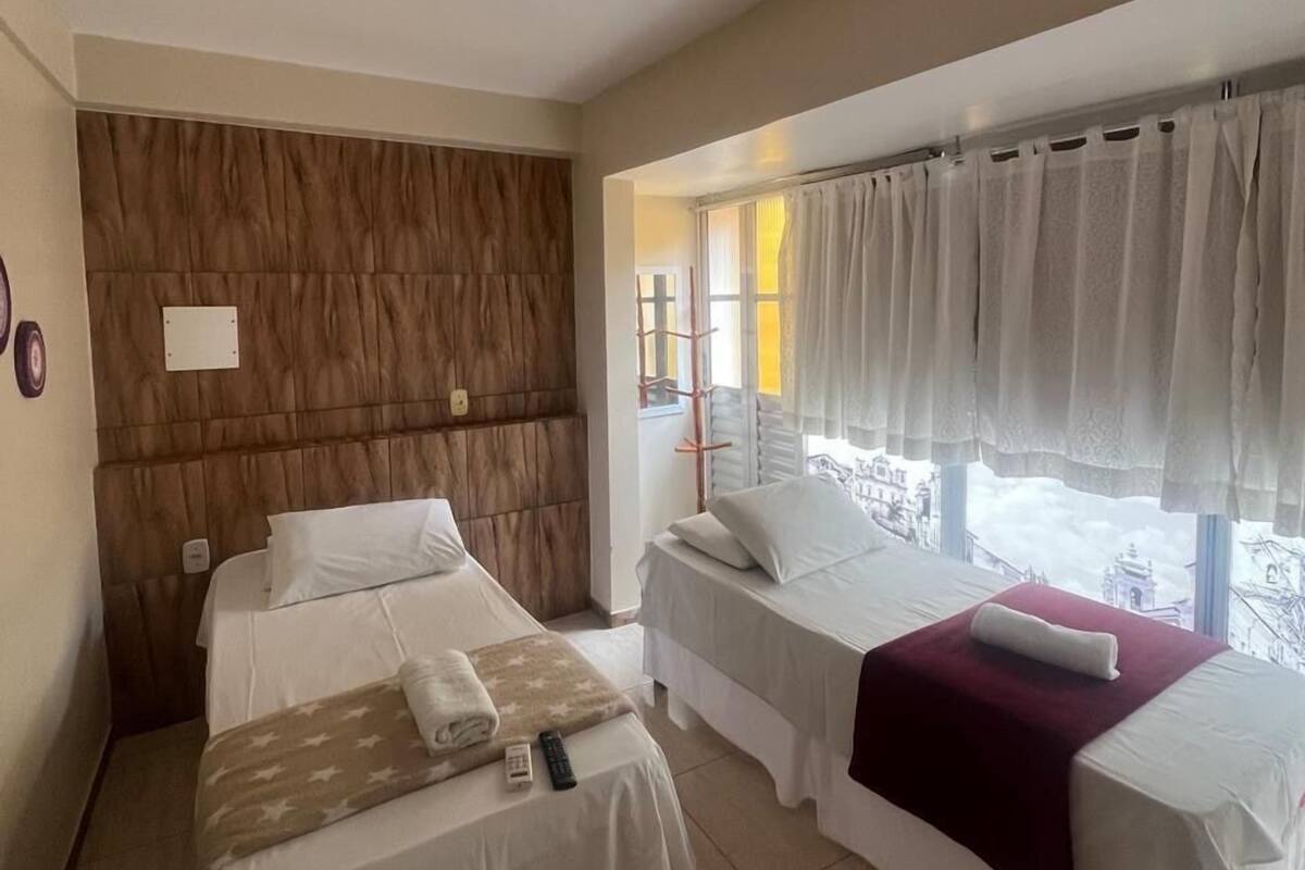 A hotel room with two beds, a wooden headboard, a window with curtains, and a view of buildings outside.