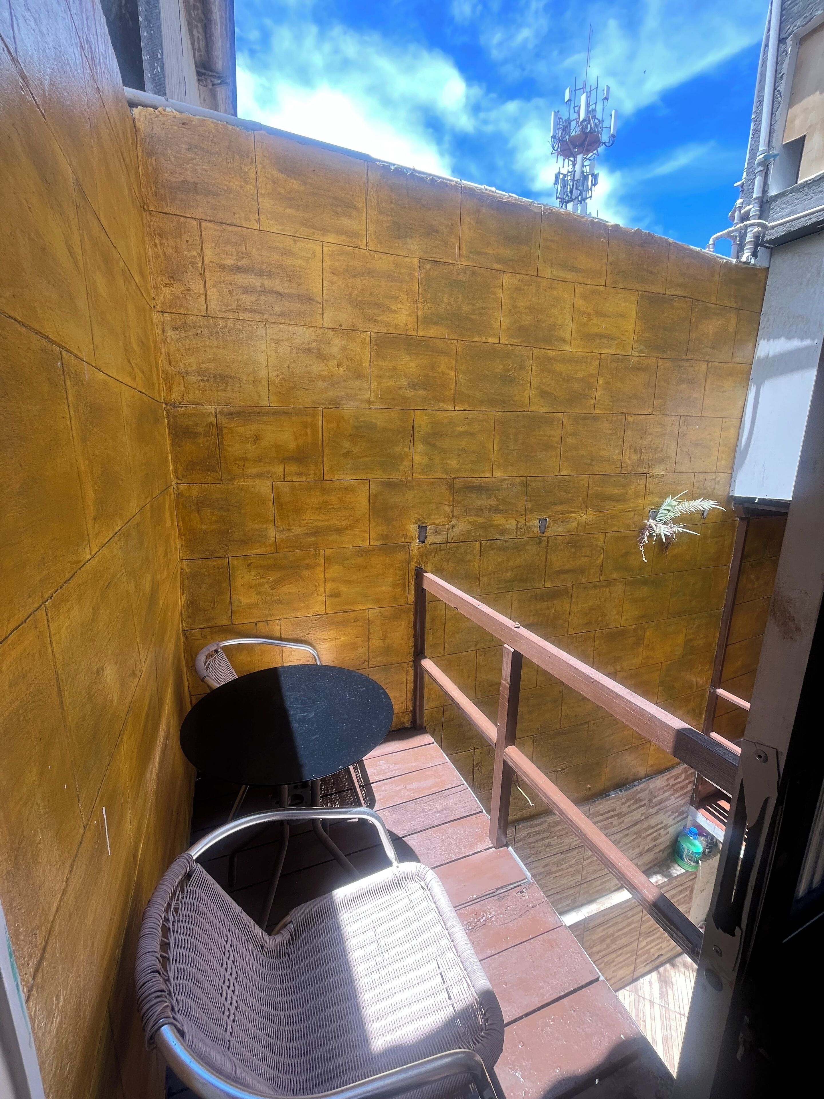 A small outdoor seating area with a wicker chair and a table, a wooden wall, and a metal railing.