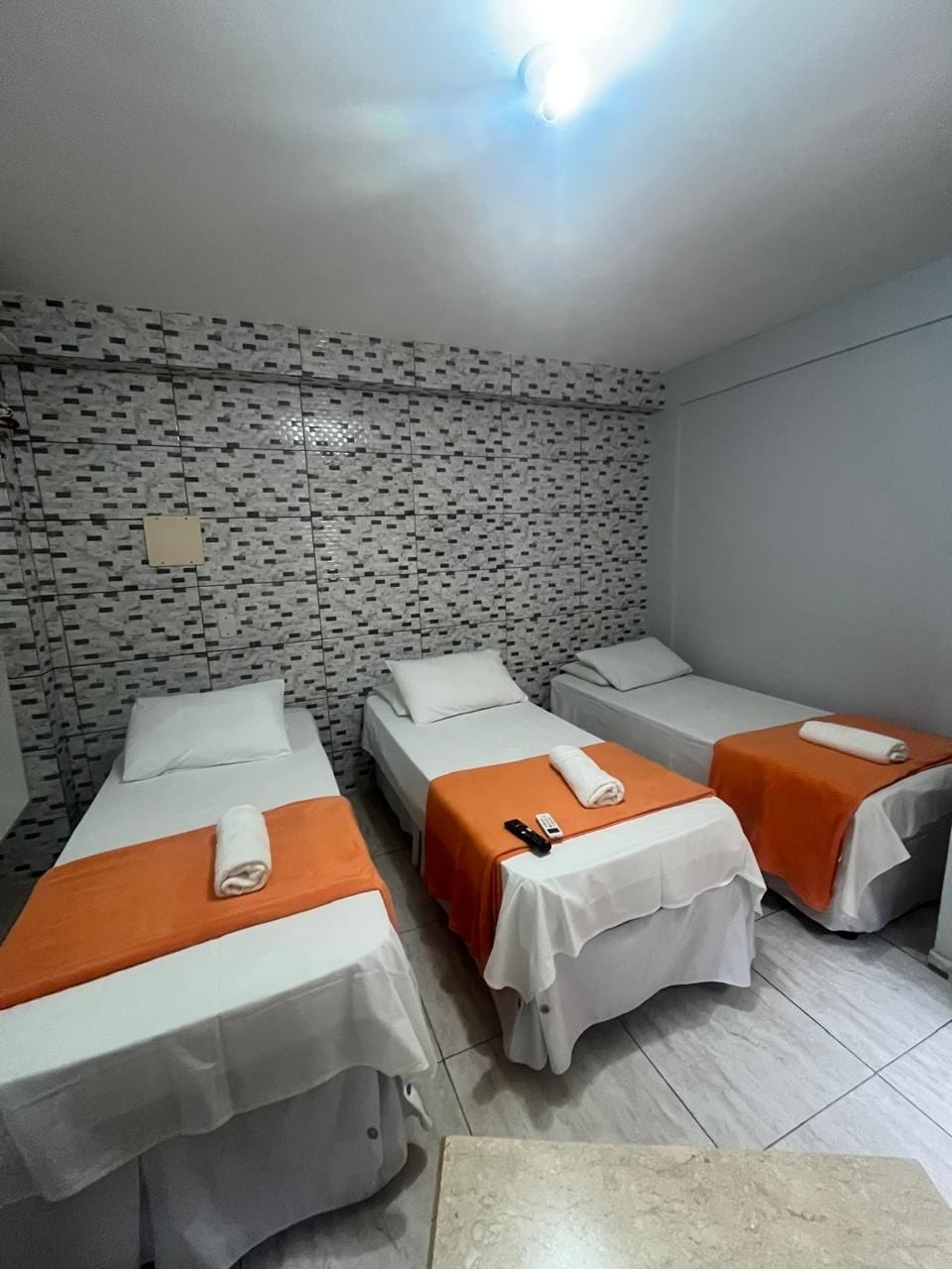 Ein Hotelzimmer mit drei Einzelbetten, jedes mit einer orangefarbenen Tagesdecke und weißer Bettwäsche. Der Raum hat eine Wand mit Muster und einen Fliesenboden.