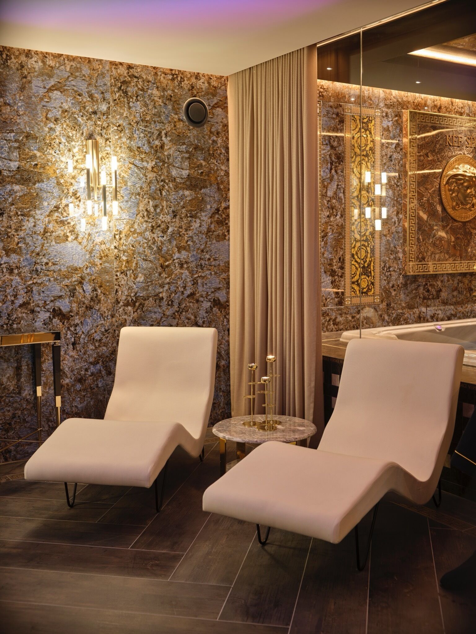 A modern bathroom with two white lounge chairs, a marble wall, a gold-framed mirror, and a gold decorative piece.