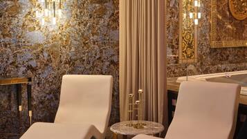 A modern bathroom with two white lounge chairs, a marble wall, a gold-framed mirror, and a gold decorative piece.