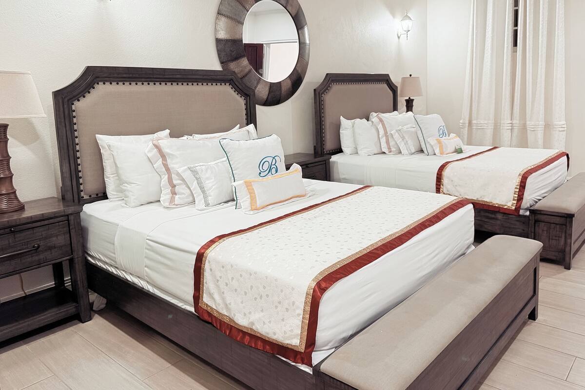 A hotel room with two beds, each with a wooden headboard, white bedding, and a decorative mirror above the headboard. There are bedside tables with lamps, a bench at the foot of the beds, and a large window with curtains.