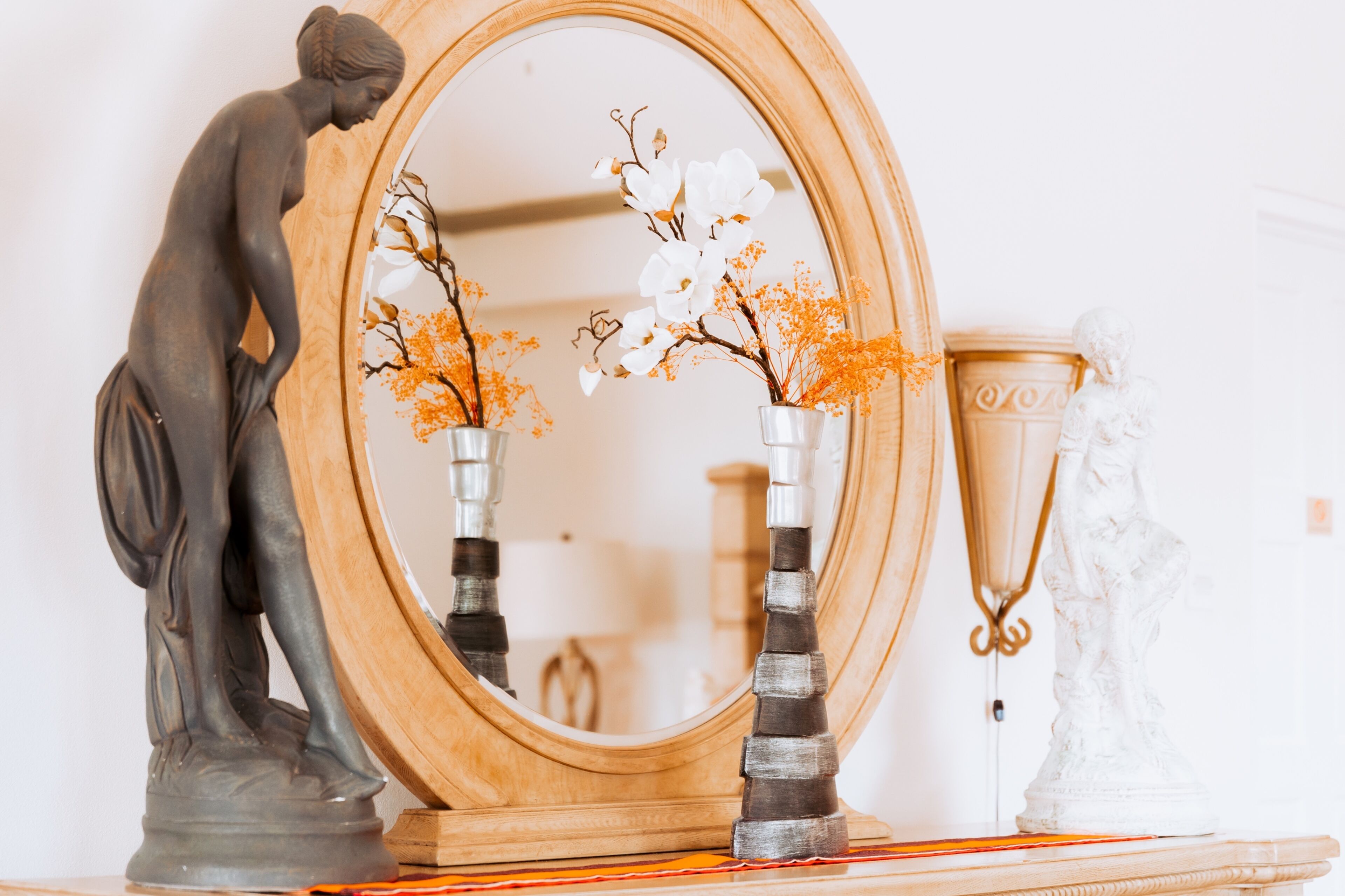 A statue of a woman, a mirror, and two vases with flowers.