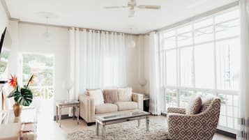 A spacious living room with a large window, a ceiling fan, a sofa, a patterned armchair, a coffee table, and a small side table with a lamp.