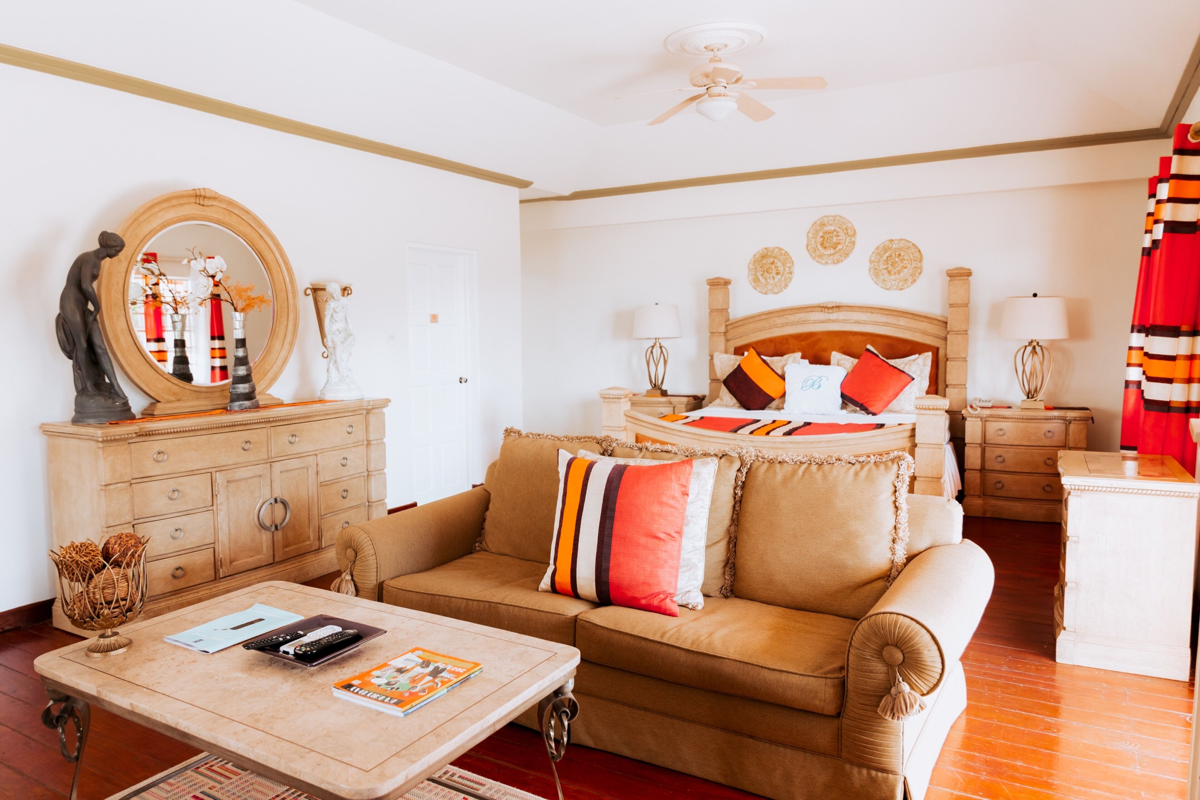 A well-lit living room with a sofa, coffee table, and a wooden cabinet. There is a ceiling fan, a bed with a wooden headboard, and a red curtain.