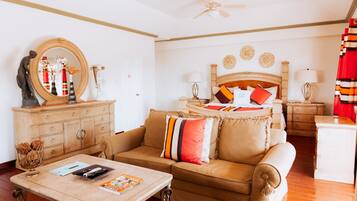 A well-lit living room with a sofa, coffee table, and a wooden cabinet. There is a ceiling fan, a bed with a wooden headboard, and a red curtain.