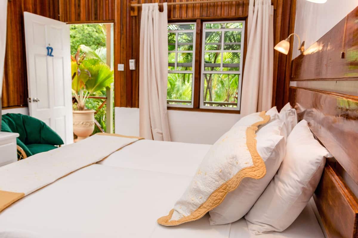 A neatly made bed with white linens and decorative pillows, a wooden headboard, and a view of lush greenery through the window.