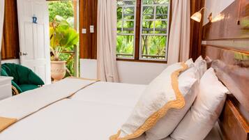 A neatly made bed with white linens and decorative pillows, a wooden headboard, and a view of lush greenery through the window.