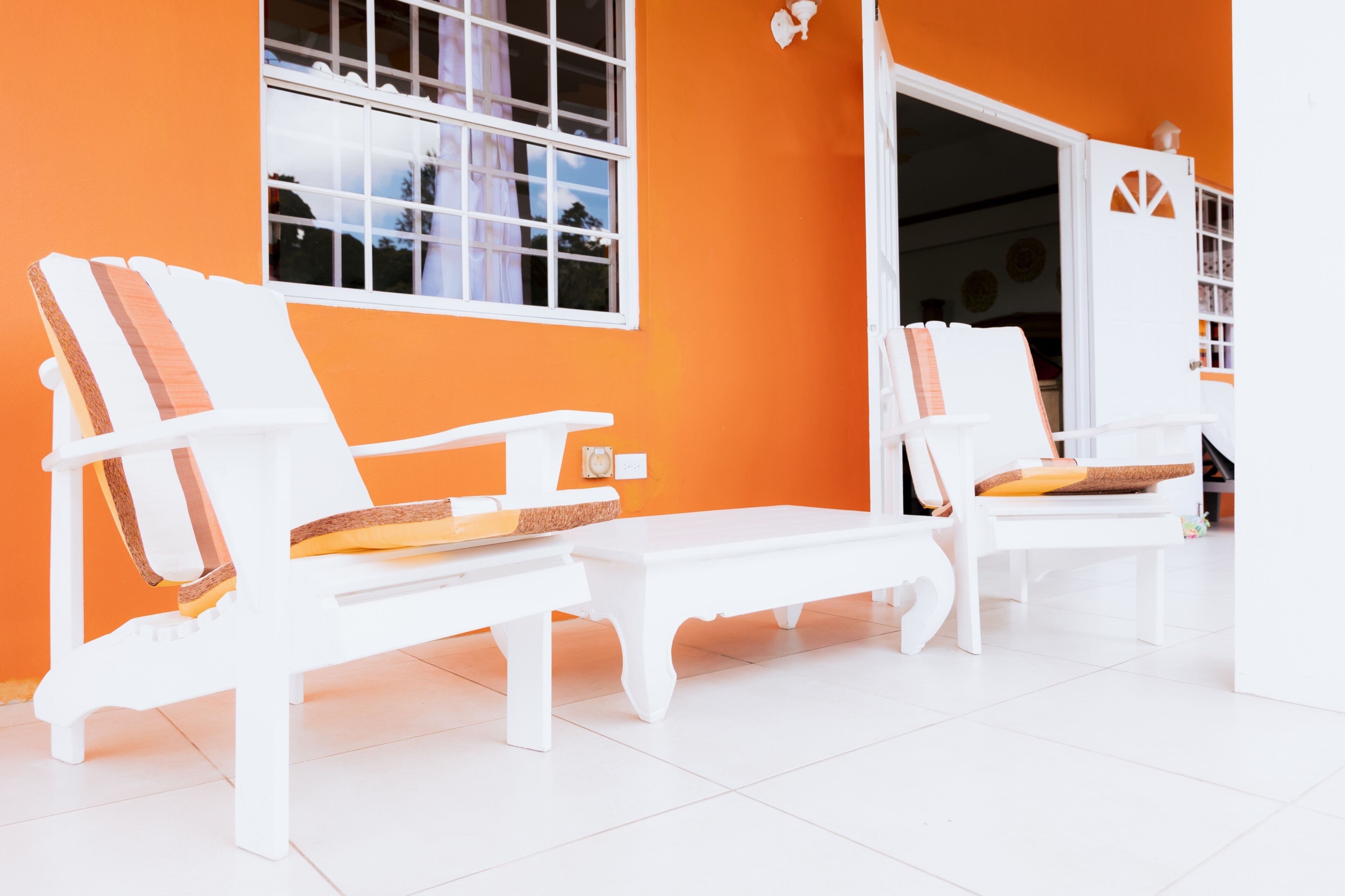Two white lounge chairs with cushions against an orange wall, a white bench, and a white door with a window.