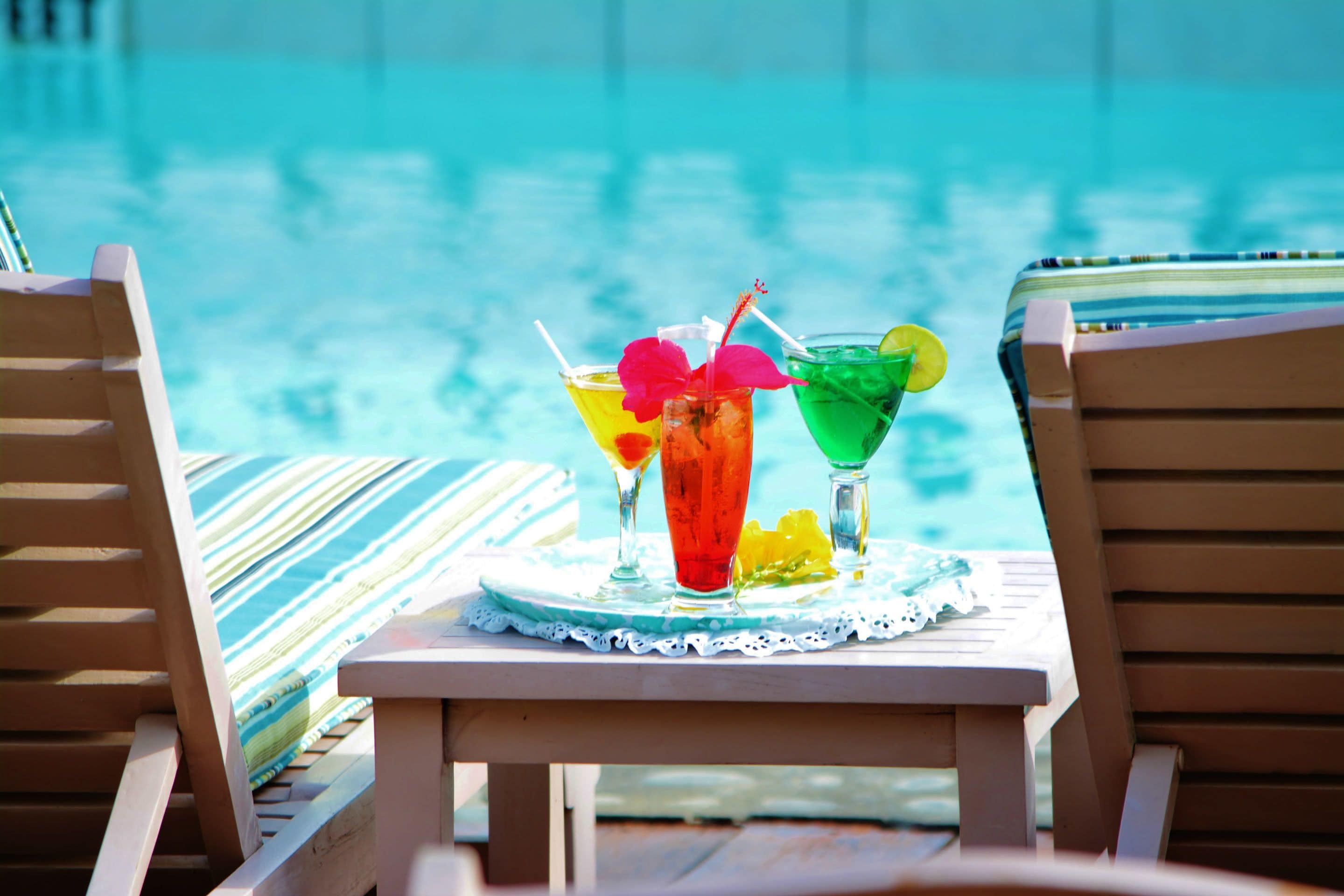 A poolside setting with two wooden lounge chairs and a table with two colorful cocktails.