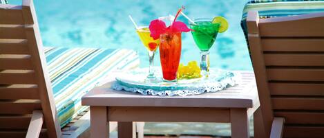 A poolside setting with two wooden lounge chairs and a table with two colorful cocktails.