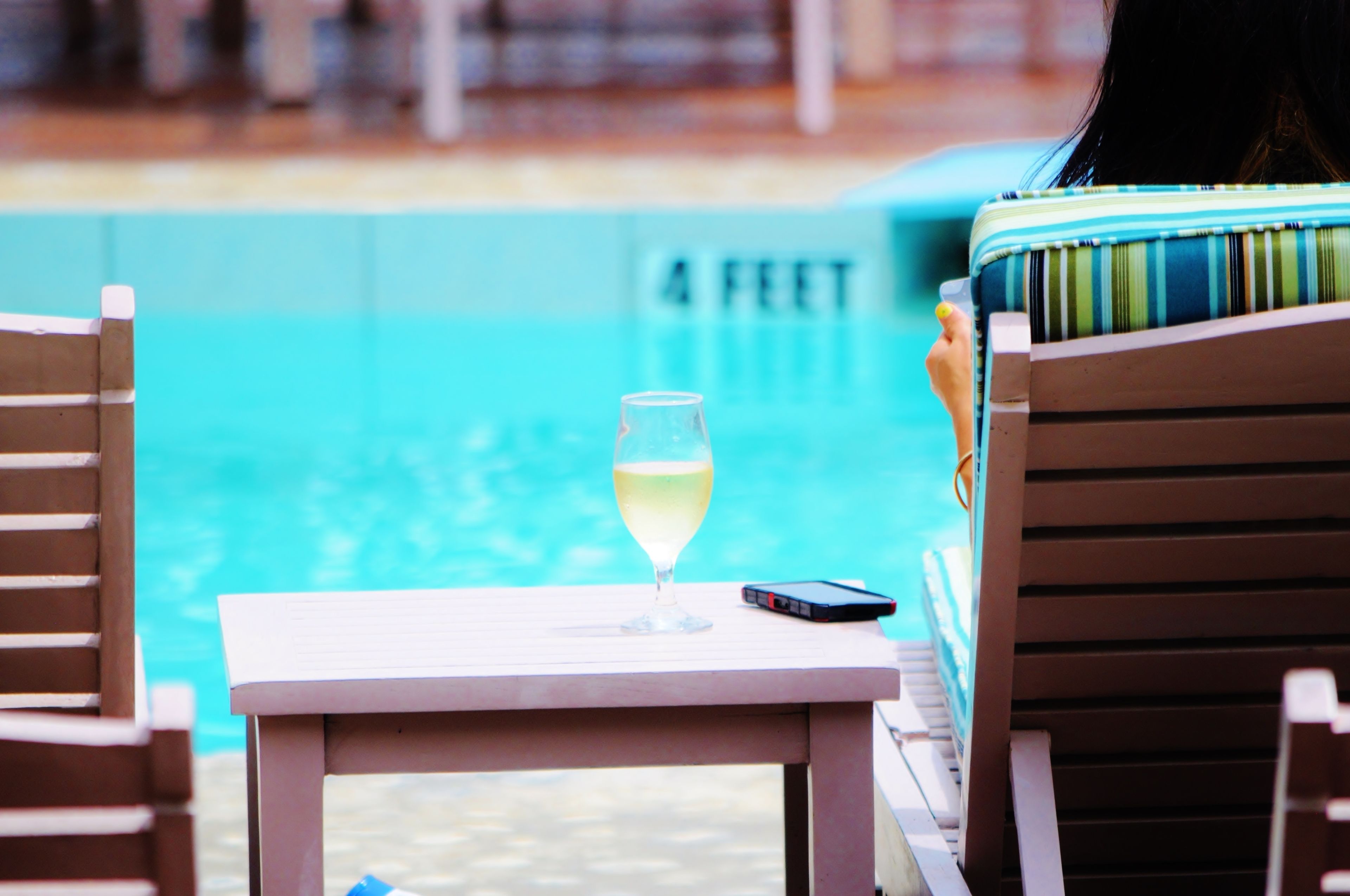 A poolside setting with a glass of white wine on a table, a smartphone, and a pool with a depth marker of 4 feet.