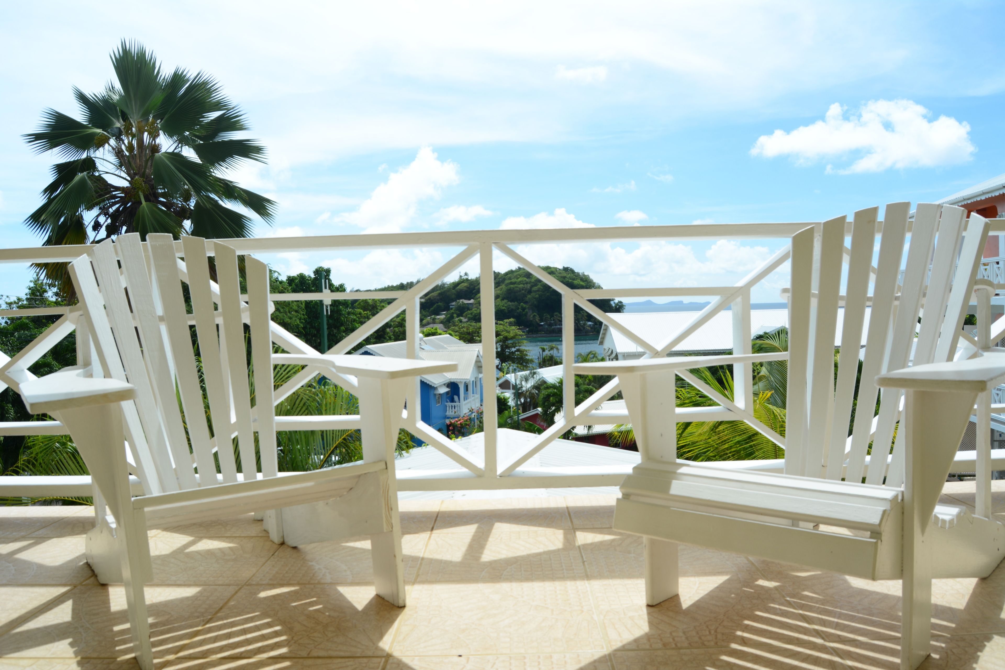 Two white Adirondack chairs on a balcony with a view of a coastal landscape and a palm tree.