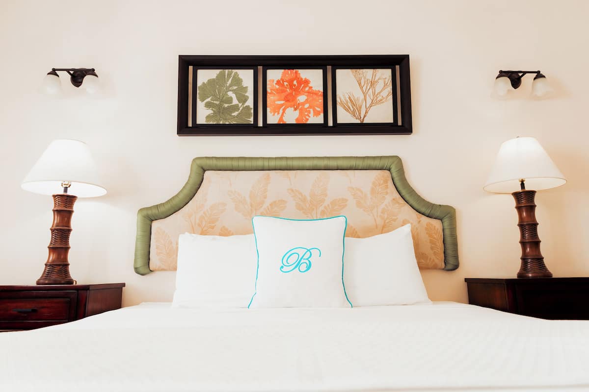 A neatly made bed with a green upholstered headboard, two bedside lamps, and a framed botanical artwork above.