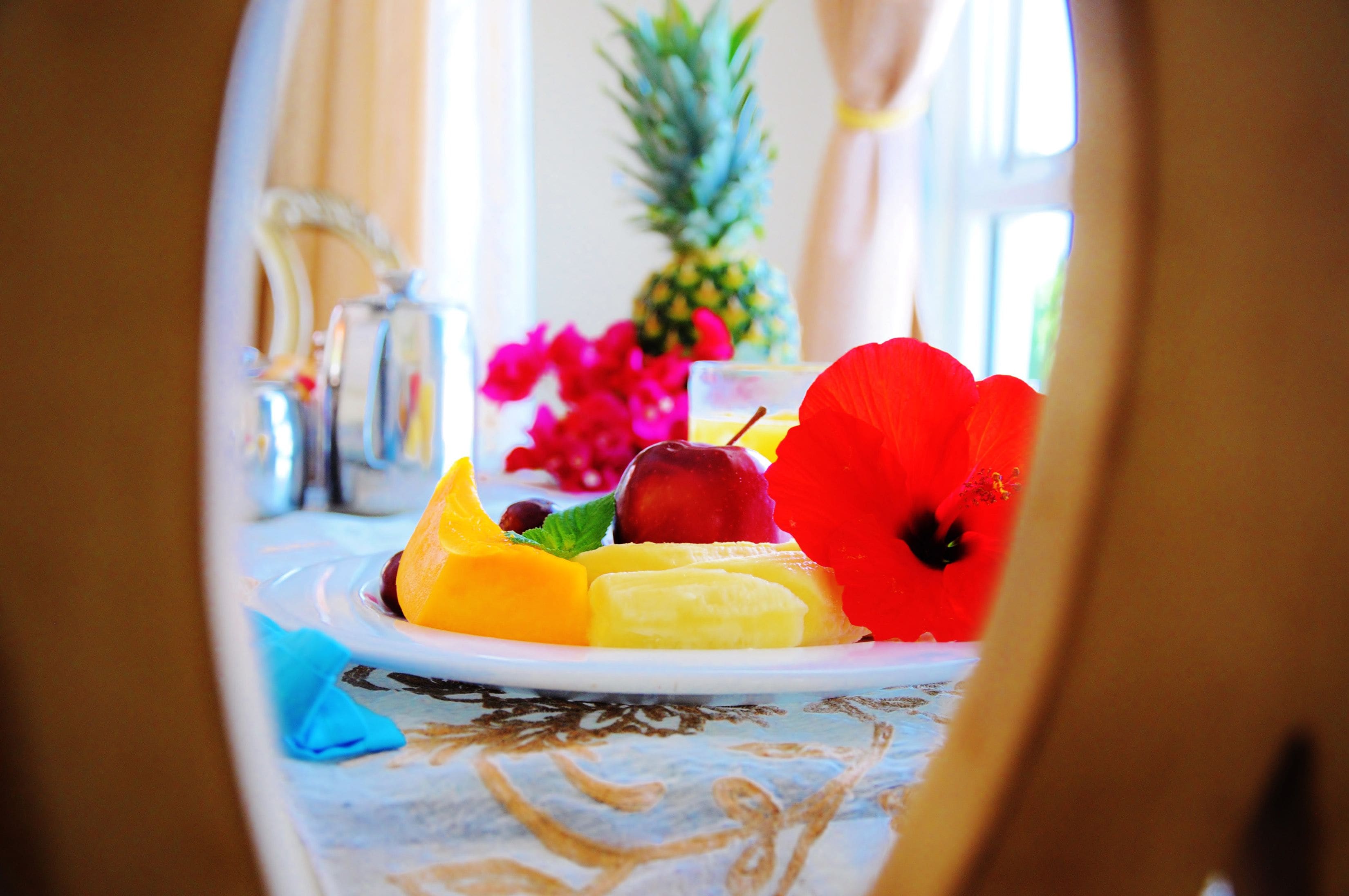 A plate of fruit with a red flower, a pineapple, and a glass of orange juice on a table with a floral tablecloth.