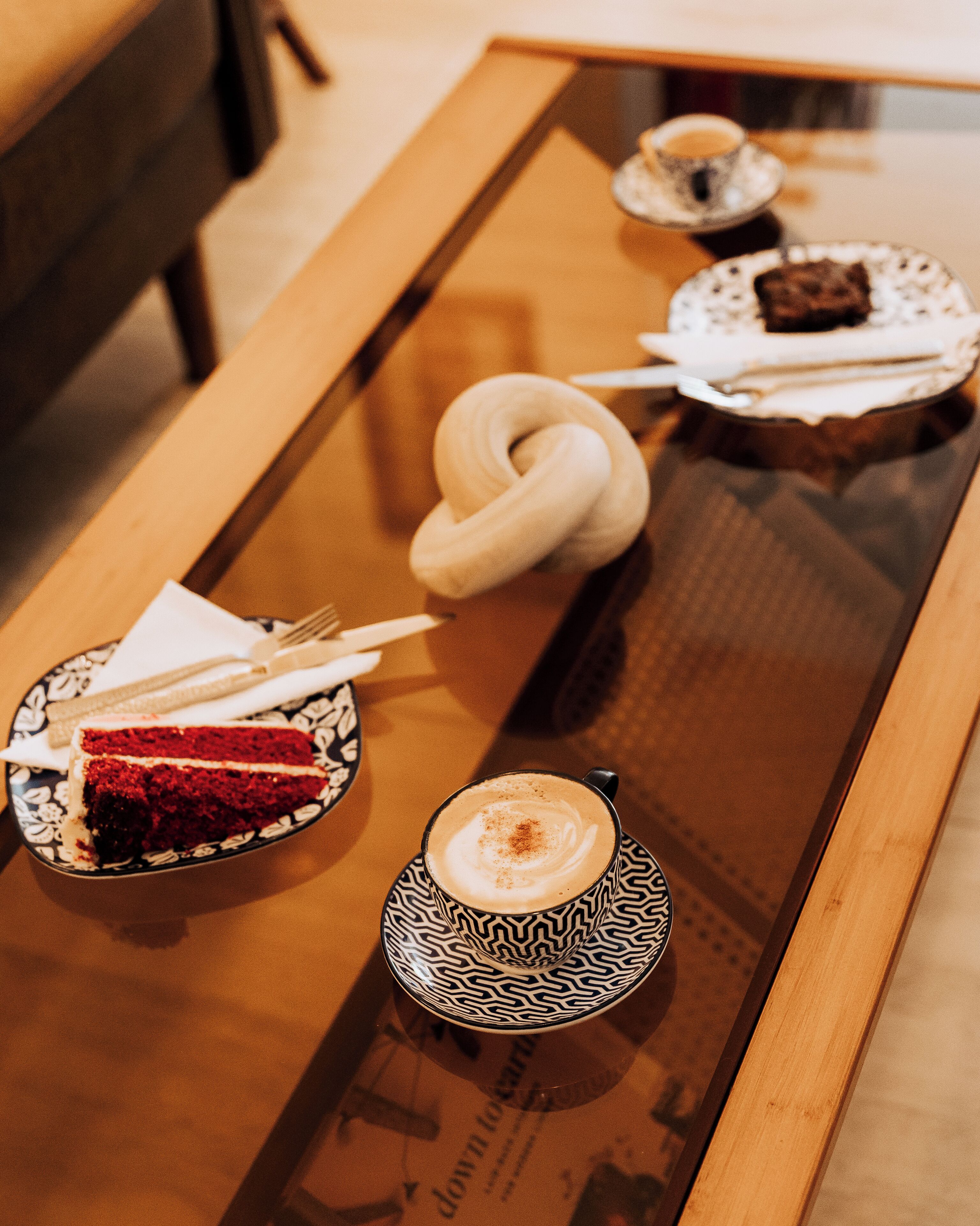 Una bandeja con una rebanada de pastel de terciopelo rojo, un pastel con forma de corazón, una taza de café con arte latte y un plato pequeño con un trozo de pastel sobre una mesa de madera.
