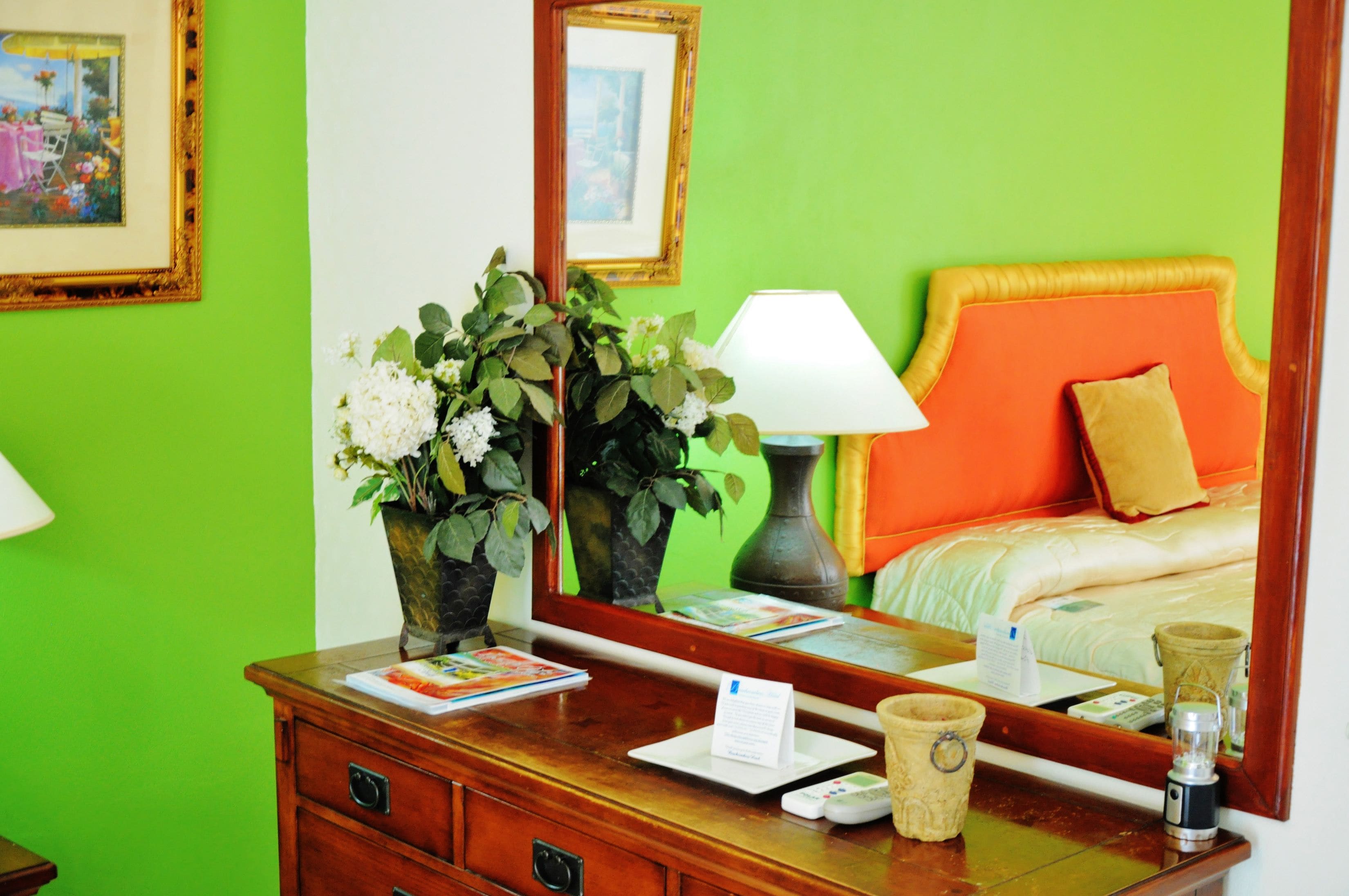 Hotelkamer met een groene wand, een houten dressoir met een bloevas, een lamp, een spiegel die een bed met een oranje hoofdeinde weerspiegelt, en een nachtkastje met een afstandsbediening en een glas.