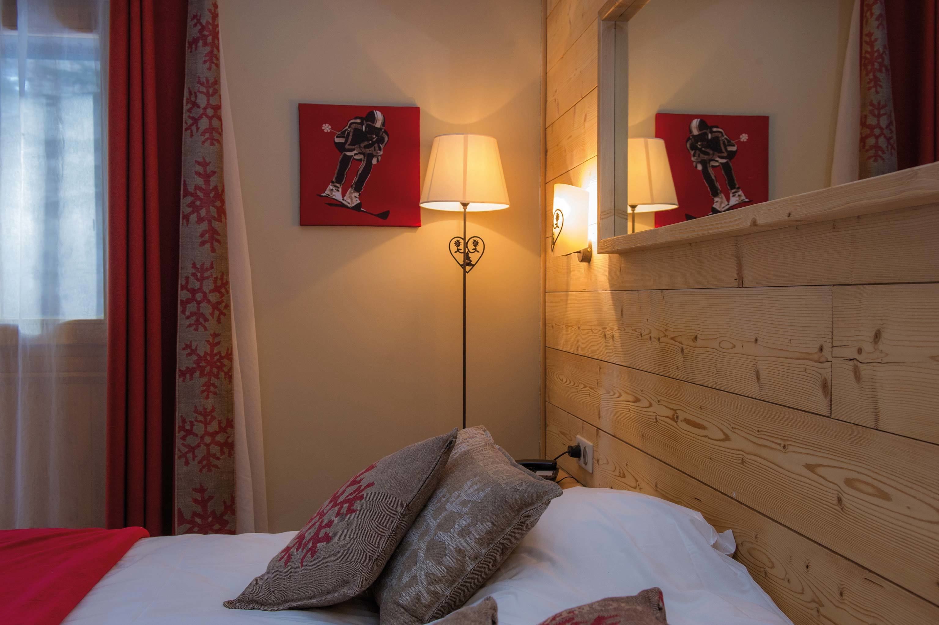 A hotel room with a wooden headboard, a bed with white linens and patterned pillows, a wall-mounted lamp, and two red artwork pieces depicting a skier.