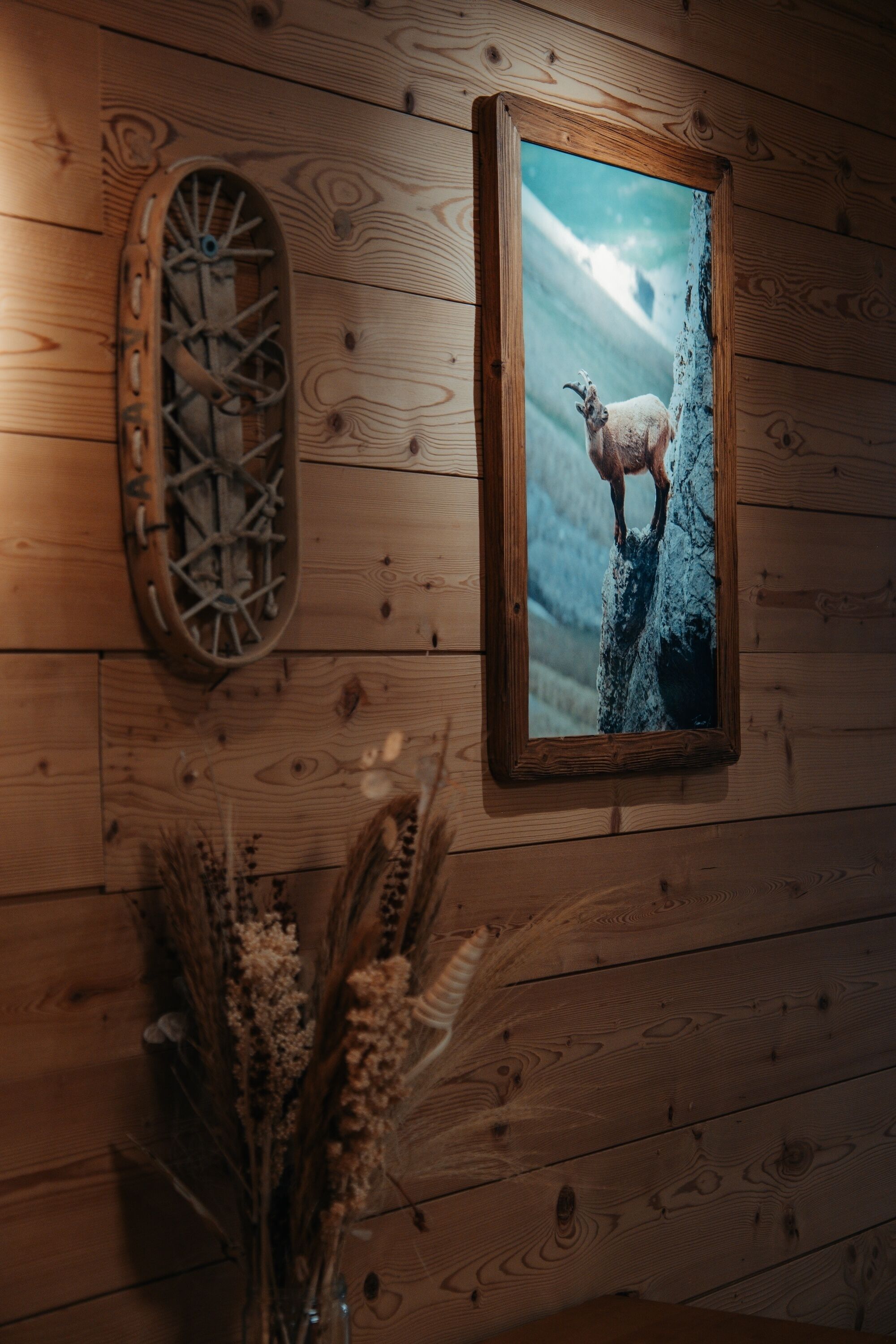 Een houten wand met een lijst waarop een geit op een rots staat, een houten wiel en gedroogde planten.