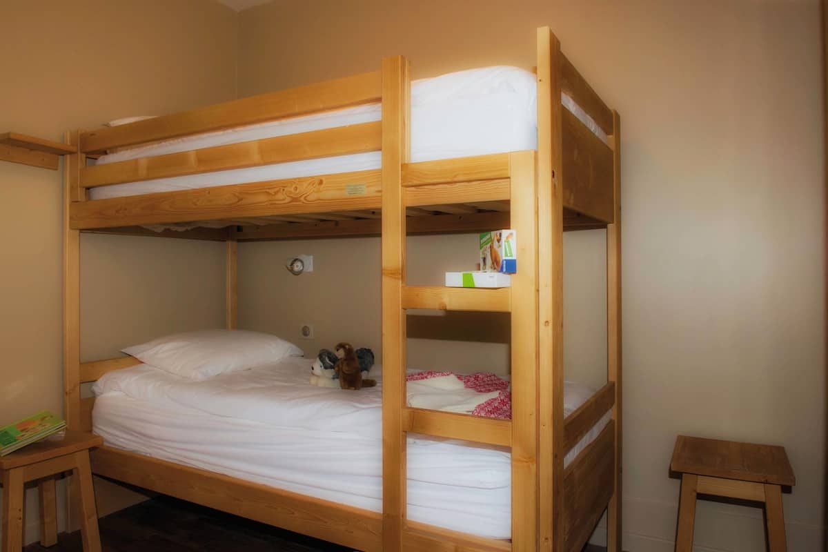 Ein Etagenbett mit einem unterem Bett und einem Regal darüber. Rechts neben dem Bett steht ein kleiner Holzschemel.