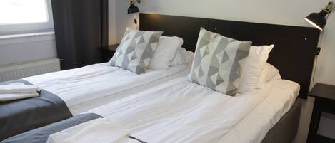 Een tweepersoonsbed met wit en grijs beddengoed, twee bedlampjes en een raam met gordijnen.