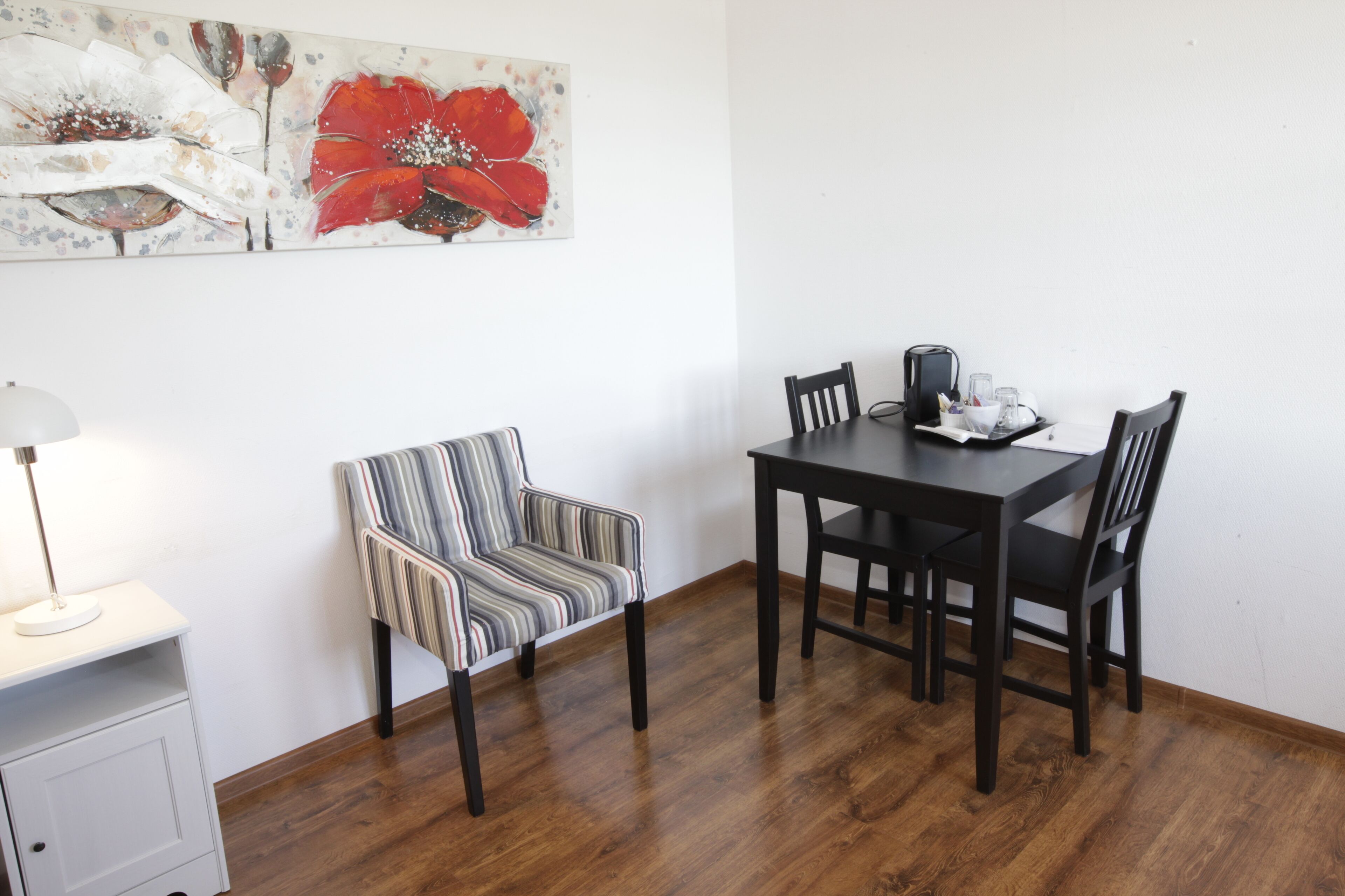A dining area with a wooden table and chairs, a striped chair, a lamp, and a painting on the wall.