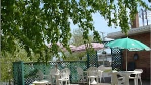 Un espace extérieur aménagé en terrasse, avec des chaises et des tables en plastique blanc, un parasol vert et, en arrière-plan, un bâtiment en briques.