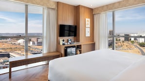 Een hotelkamer met een groot bed, een aan de muur bevestigde televisie, een klein bureau en uitzicht op de stad door grote ramen.