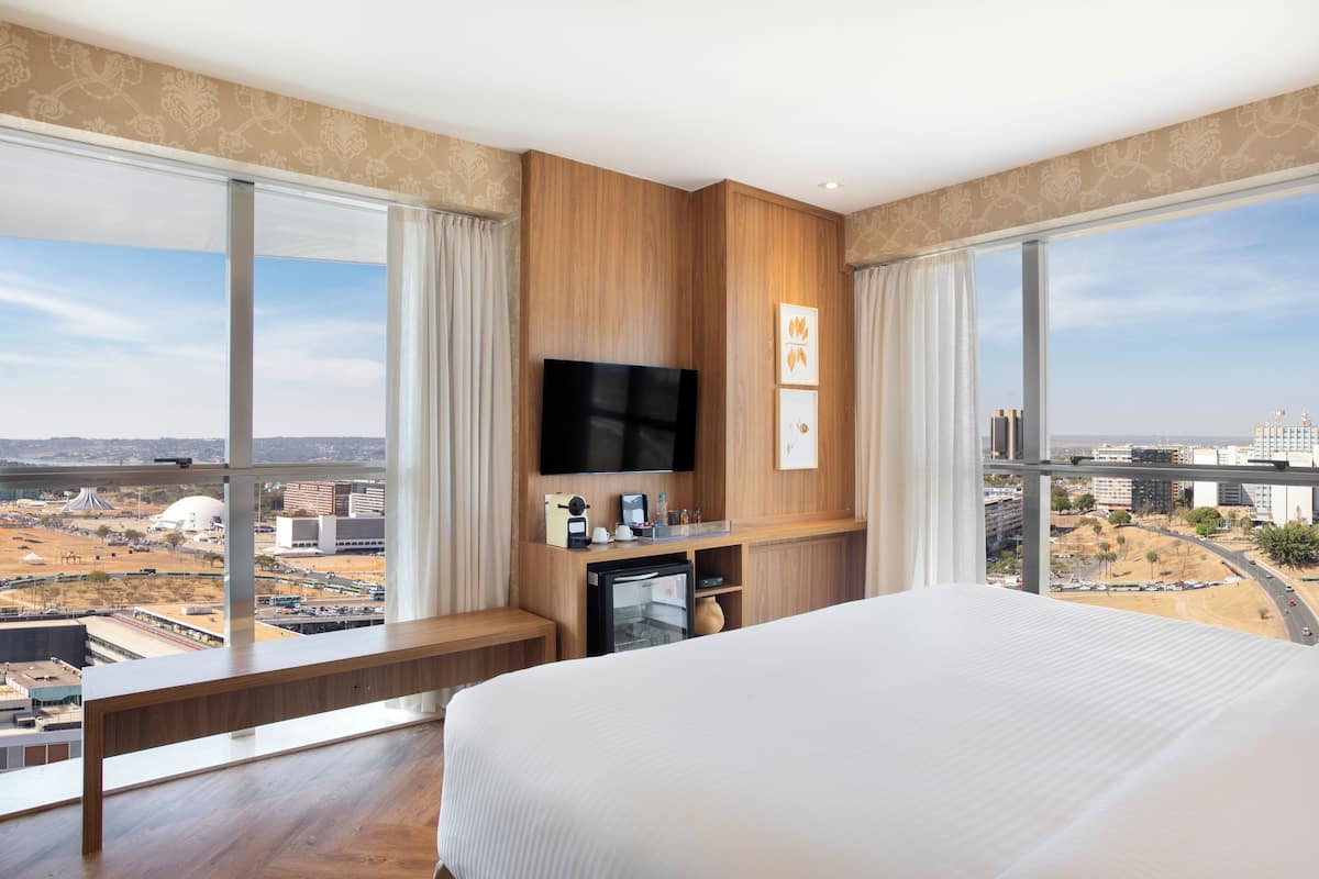 Een hotelkamer met een groot bed, een aan de muur bevestigde televisie, een klein bureau en uitzicht op de stad door grote ramen.