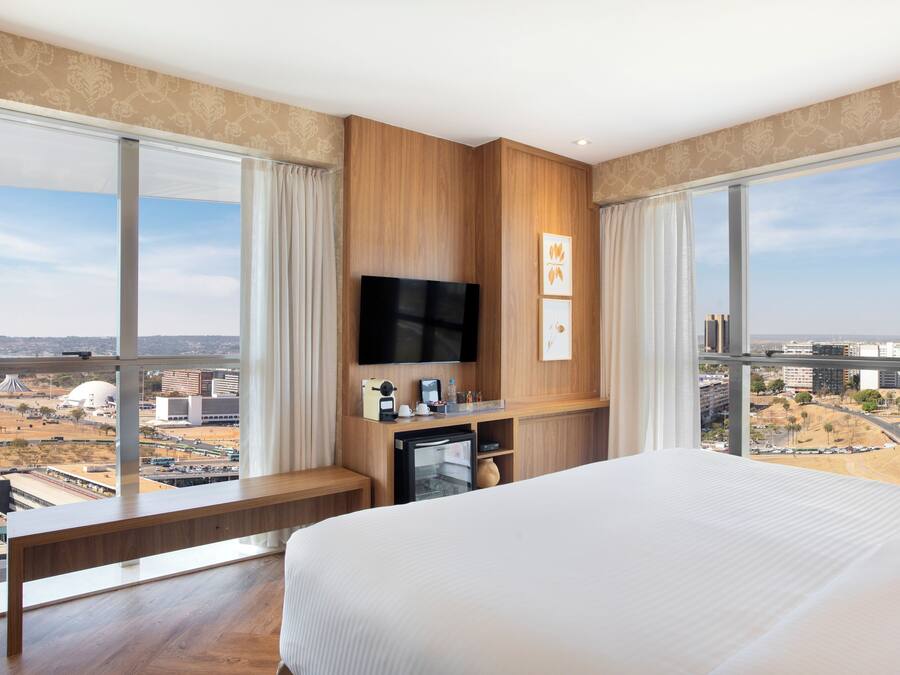 Een hotelkamer met een groot bed, een aan de muur bevestigde televisie, een klein bureau en uitzicht op de stad door grote ramen.