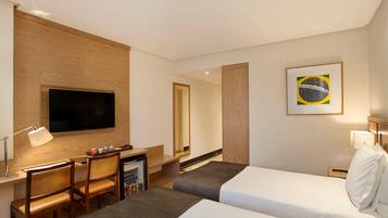 A hotel room with two beds, a desk with a chair, a television, a minibar, and a framed picture on the wall.