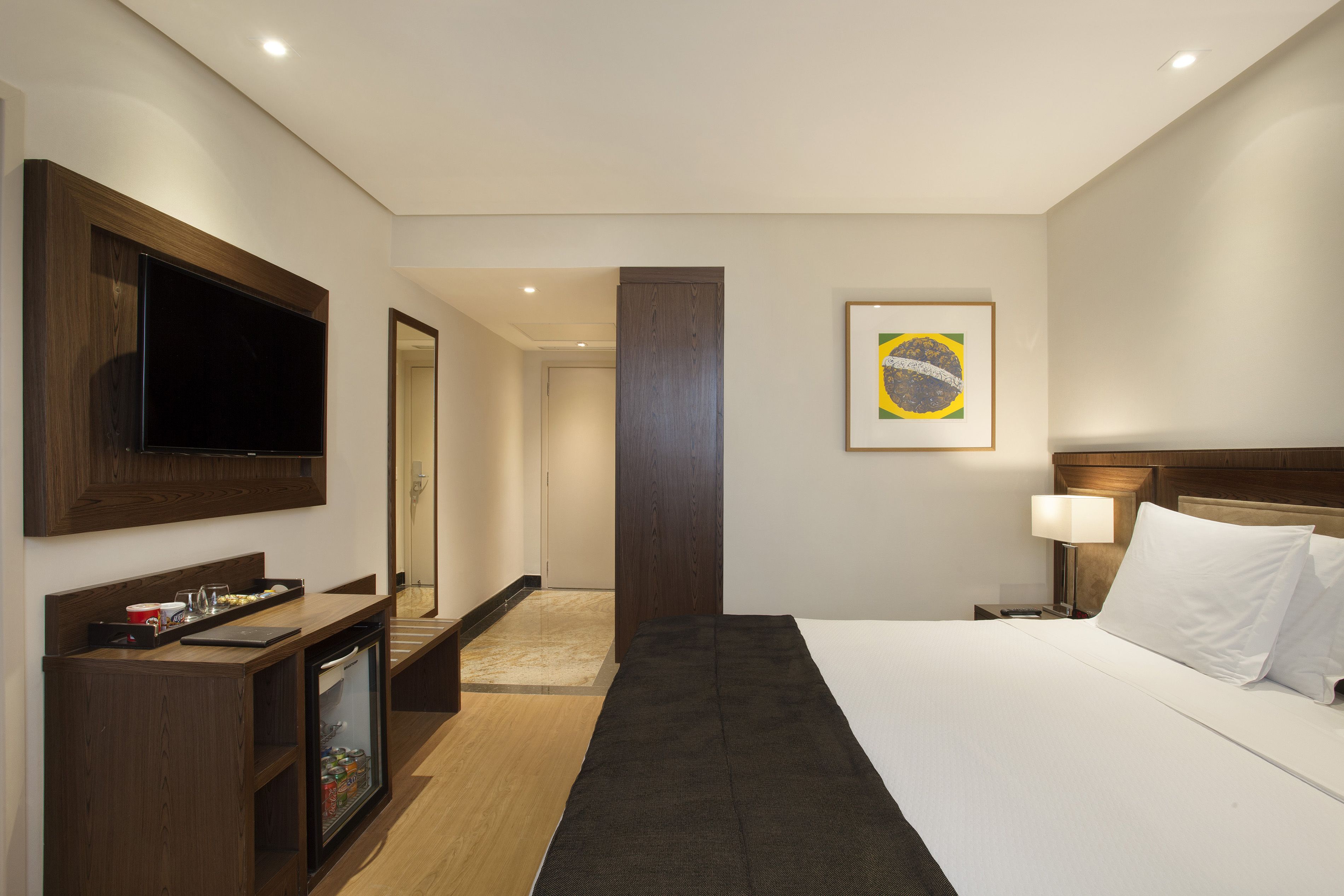 A hotel room with a bed, a TV, a minibar, a desk, and a door leading to another room.