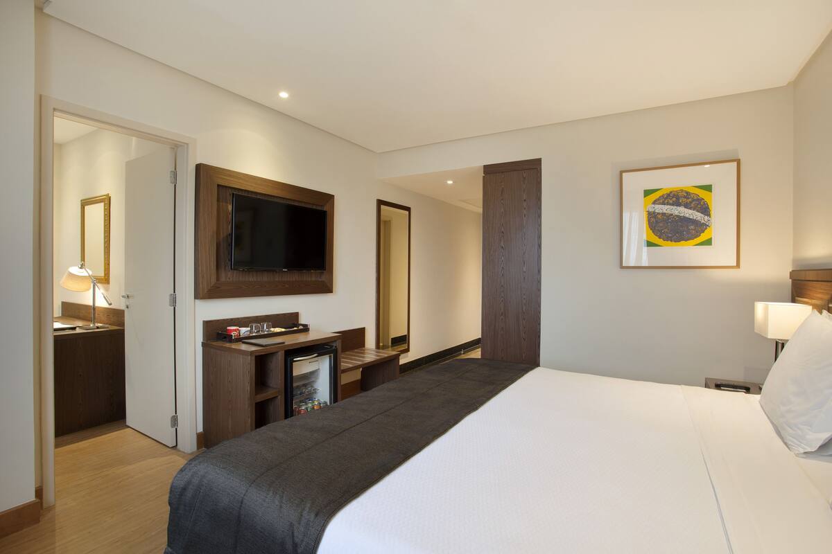 Een hotelkamer met een bed, een aan de muur bevestigde televisie, een bureau met een computer, een kleine koelkast en een deur naar de badkamer.