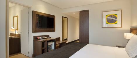 A hotel room with a bed, a TV mounted on the wall, a desk with a computer, a small fridge, and a door leading to a bathroom.