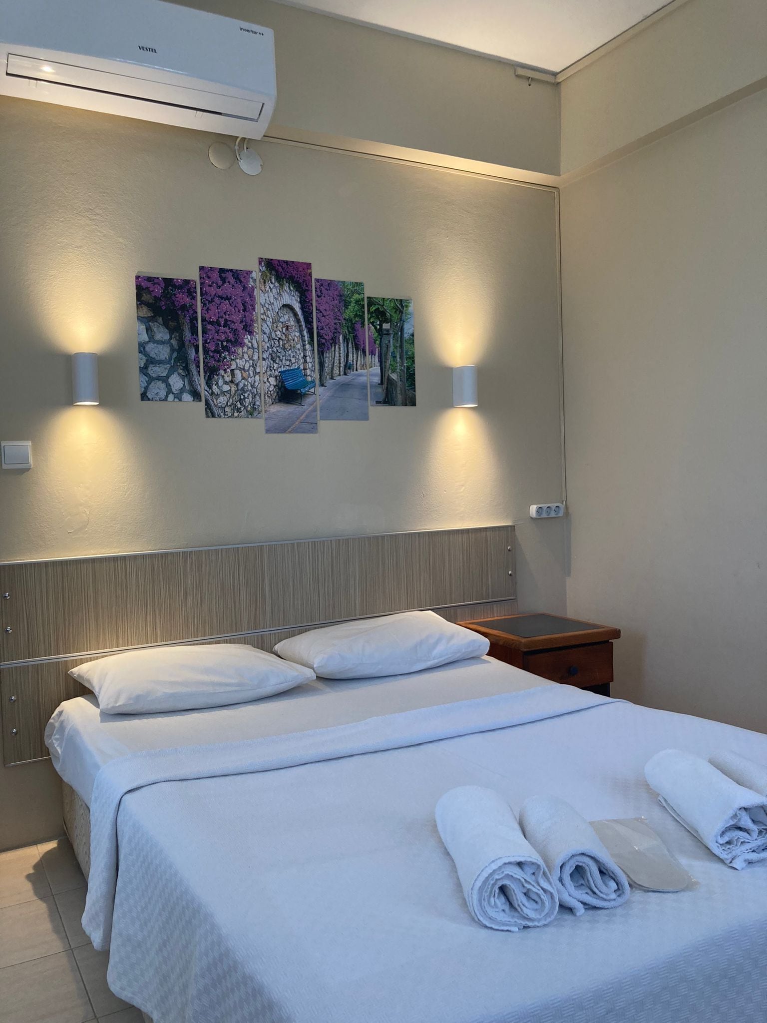 A hotel room with a bed, two wall-mounted lamps, an air conditioner, and three framed pictures on the wall.